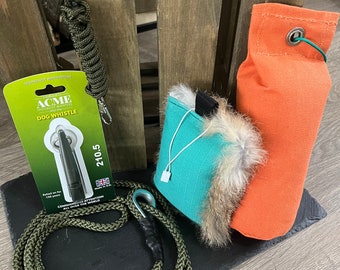 May include: Green dog training rope with a black handle, a green and turquoise dog training dummy, an orange dog training dummy, and a black and silver dog whistle with the text "ACME SPECIALIST WHISTLES DOG WHISTLE 210.5" on a white and green label.