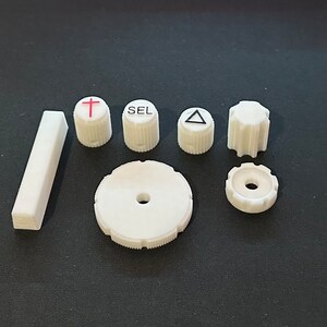 May include: A set of six white plastic knobs with various symbols, including a cross, a triangle, and the word "SEL". There is also a white plastic rectangular block.
