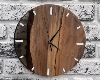 Puede incluir: Un reloj de pared redondo con una esfera de madera natural y una sección de resina oscura y translúcida en el lado izquierdo. El reloj tiene marcadores de hora rectangulares blancos y manecillas de metal negro finas en el centro.