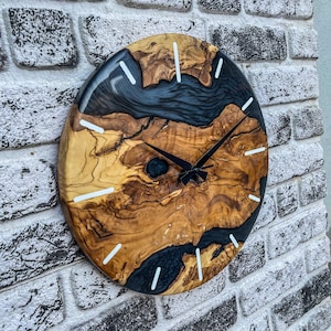 May include: A round wooden clock with black hands and white markings. The clock face is made of wood with a black epoxy resin inlay. The clock is hanging on a white brick wall.