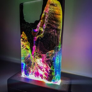 May include: A colorful resin lamp with a black base. The resin has a swirling, abstract pattern with shades of green, blue, purple, and gold. The lamp is lit from below, casting a rainbow glow.