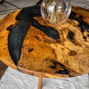 Op de afbeelding: Een ronde houten tafelblad met een zwarte epoxyhars inlay. Het tafelblad heeft een natuurlijke houtnerf en is afgewerkt met een transparante laag. Het tafelblad is ongeveer 61 cm in diameter.