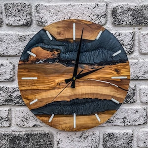 Puede incluir: Un reloj redondo de madera con un diseño de resina epoxi negra y gris. El reloj tiene marcadores de hora blancos y manecillas negras.