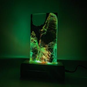 May include: A green and gold resin lamp with a wooden base. The lamp is lit and casting a green glow.
