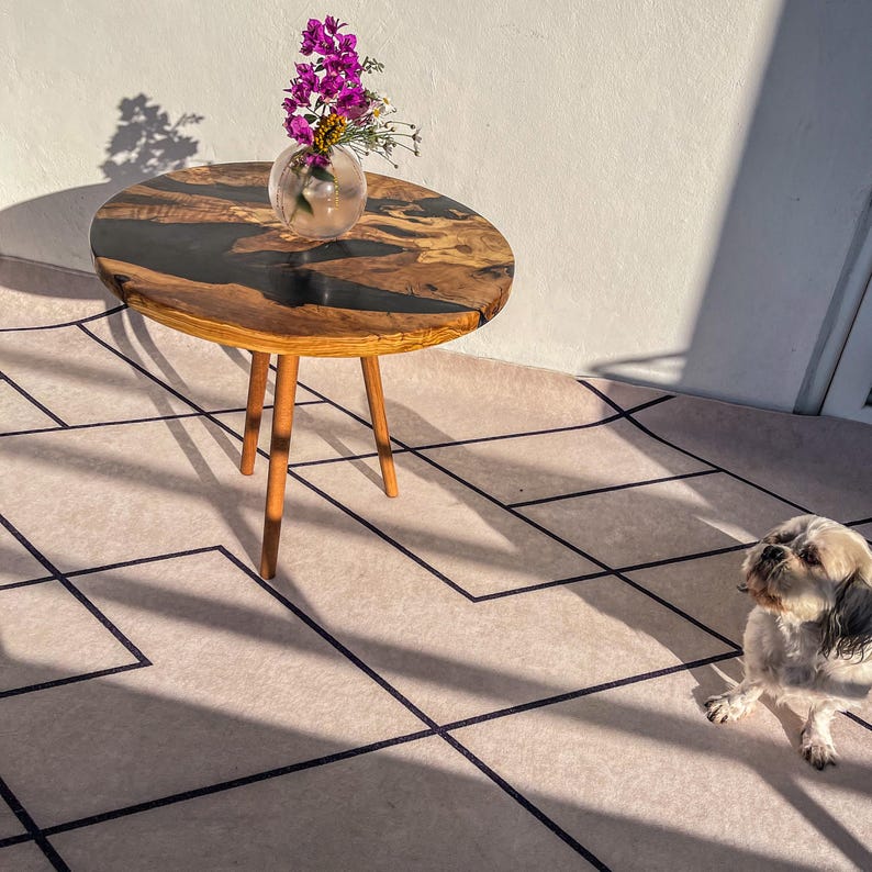 Op de afbeelding: Een kleine ronde houten tafel met een zwart epoxyhars blad. De tafel heeft drie houten poten en staat op een beigekleurig tapijt met zwarte lijnen. Een vaas met paarse bloemen staat op de tafel.