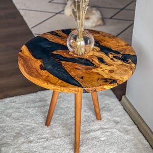 Op de afbeelding: Een ronde houten bijzettafel met een zwarte epoxyhars inlay. De tafel heeft drie houten poten en staat op een wit tapijt.