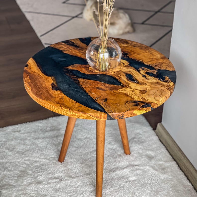 Op de afbeelding: Een ronde houten bijzettafel met een zwarte epoxyhars inlay. De tafel heeft drie houten poten en staat op een wit tapijt.