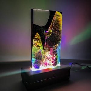 May include: A colorful resin lamp with a black base. The resin is translucent and has a swirling pattern of green, yellow, and pink. The lamp is lit and casting a rainbow glow.