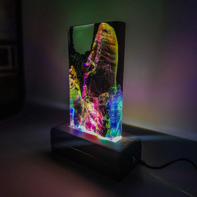 May include: A colorful resin lamp with a wooden base. The resin is translucent and has a swirling pattern of rainbow colors. The lamp is lit and casting a warm glow.