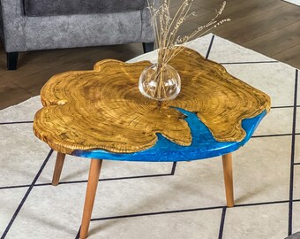 May include: A distinctive coffee table featuring a live edge wooden top with a blue resin river design. The table has a natural wood grain and is supported by three wooden legs. A small glass vase with dried plants is placed on the table.