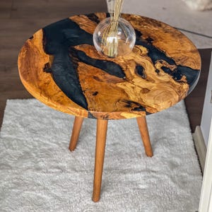 Op de afbeelding: Een ronde houten tafel met een zwarte epoxyhars inlay. De tafel heeft drie houten poten en staat op een wit tapijt.