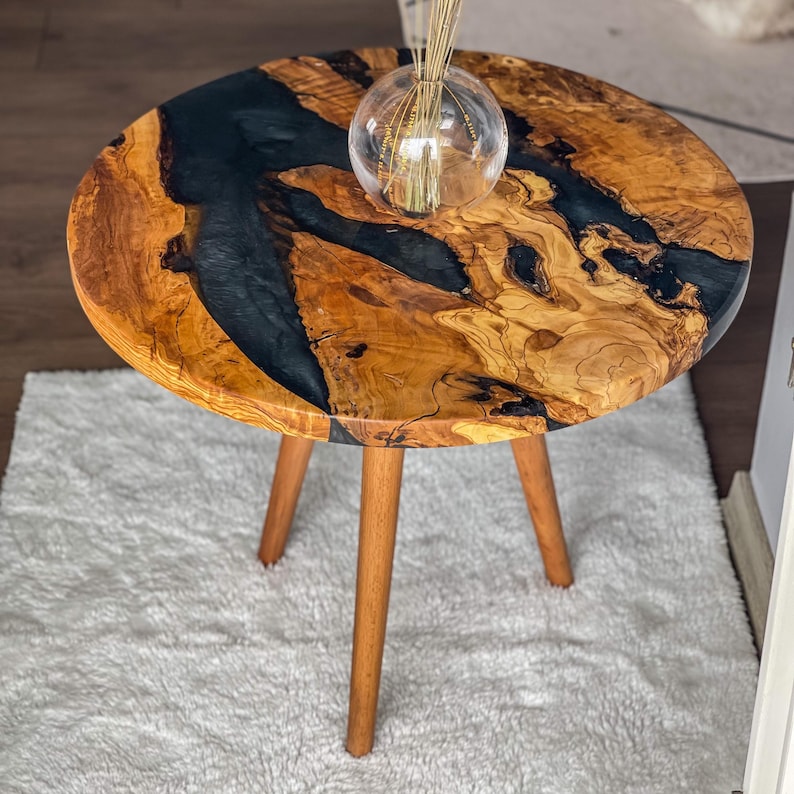 Op de afbeelding: Een ronde houten tafel met een zwarte epoxyhars inlay. De tafel heeft drie houten poten en staat op een wit tapijt.