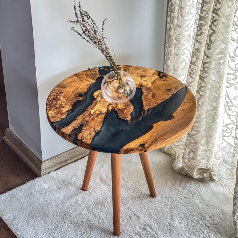 Op de afbeelding: Een ronde houten bijzettafel met een zwarte epoxyhars inlay. De tafel heeft drie houten poten en staat op een wit tapijt. Een glazen vaas met gedroogde bloemen staat op de tafel.