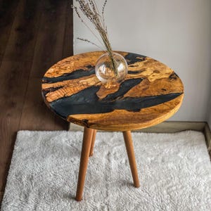 Op de afbeelding: Een ronde houten bijzettafel met een zwarte epoxyhars inlay. De tafel heeft drie houten poten en staat op een wit tapijt.