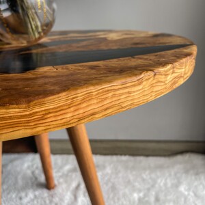 Op de afbeelding: Een ronde houten tafel met een zwarte epoxyhars inlay. De tafel heeft drie houten poten en een natuurlijke houten afwerking.