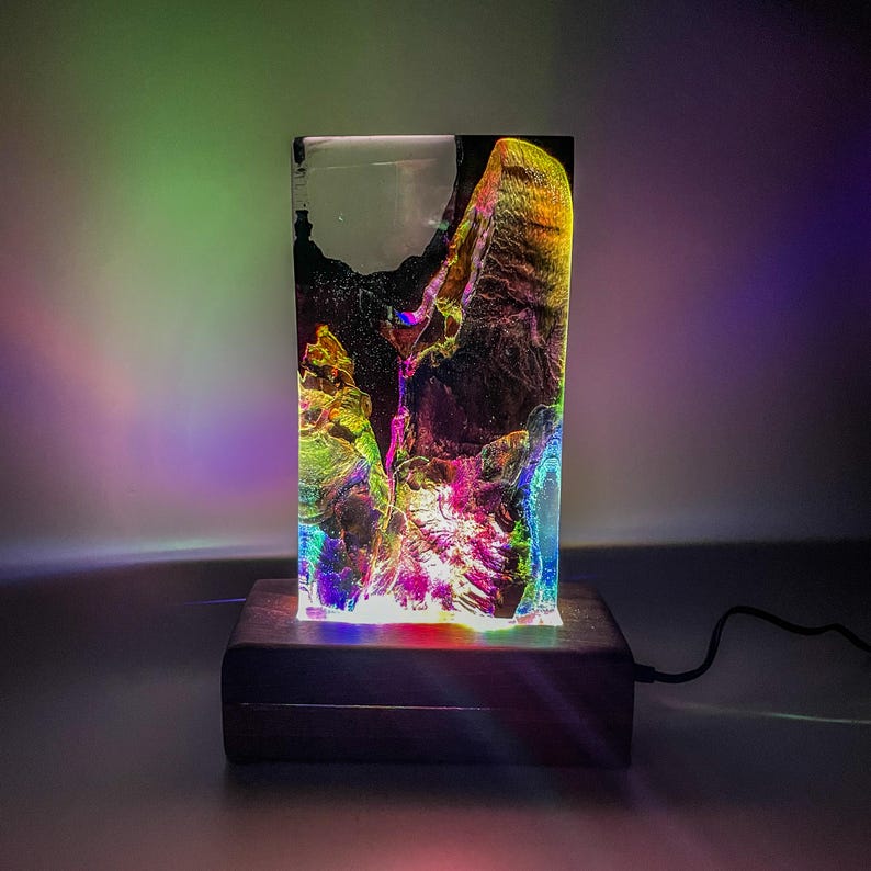 May include: A colorful resin lamp with a wooden base. The lamp is lit and casts a rainbow glow.