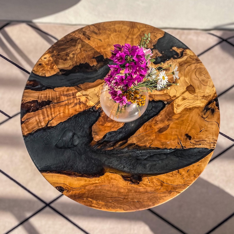 Op de afbeelding: Een ronde houten tafelblad met een zwarte epoxyhars inlay. Het tafelblad heeft een natuurlijke houtnerf en is versierd met een vaas met roze bloemen.