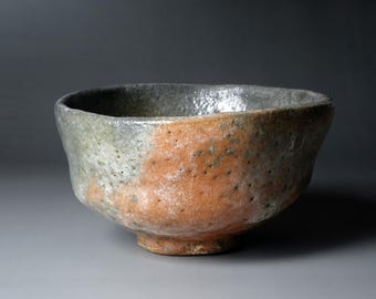 May include: A handcrafted ceramic bowl with a rustic aesthetic. The bowl features a rounded shape with a footed base. The exterior is a blend of gray and orange-brown hues, with a textured surface. The bowl is suitable for serving or display.