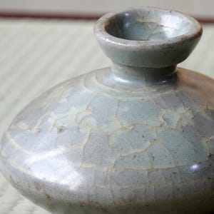 Könnte beinhalten: Nahaufnahme eines keramischen Gefäßes in Celadon-Farbe mit bauchigem Körper und schmalem Hals. Die Oberfläche weist einen Craquelé-Effekt auf, der Textur verleiht. Das Gefäß dient wahrscheinlich dekorativen Zwecken.