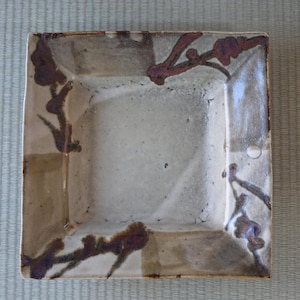 Puede incluir: Plato cuadrado de cerámica con un centro beige claro y un borde más oscuro y moteado. El plato presenta diseños marrones en forma de rama en cada esquina, añadiendo un toque orgánico. La estética general es rústica y hecha a mano.