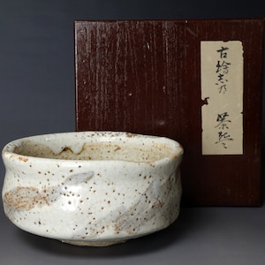 May include: A white ceramic tea bowl with brown speckles and a subtle glaze. The bowl is sitting on a grey surface next to a wooden box with a label that reads "石  茶  碗  古  美術".