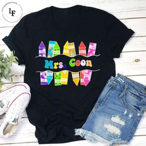 May include: Black t-shirt featuring a design of colourful crayons and the text "Mrs. Coon" in a white banner.