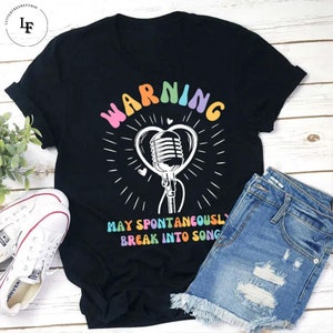 Puede incluir: Camiseta negra con un micrófono blanco dentro de un corazón y el texto "WARNING MAY SPONTANEOUSLY BREAK INTO SONG" en colores arcoíris.