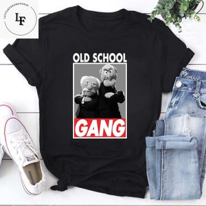 May include: Black t-shirt featuring a graphic of two puppet characters with the text "OLD SCHOOL GANG" in a white box. The shirt is paired with blue jeans and white sneakers.