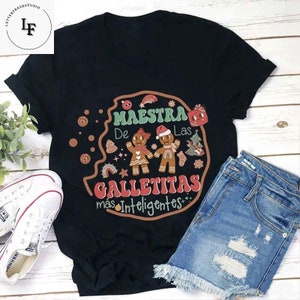 May include: Black t-shirt featuring a graphic of two gingerbread people and the text "Maestra De Las Galletitas Mas Inteligentes".