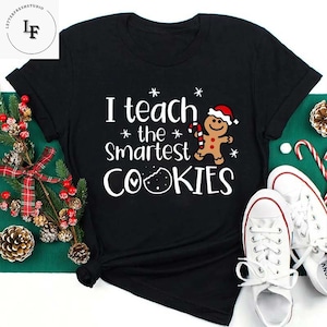 May include: Black t-shirt with white text that reads "I teach the smartest cookies". A gingerbread man with a red Santa hat and candy cane is holding a cookie.