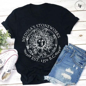 May include: Black t-shirt with a white graphic of Medusa's Stoneworks, a circular design with a stylized image of Medusa's head and the text "Medusa's Stoneworks Est. 1350 B.C."
