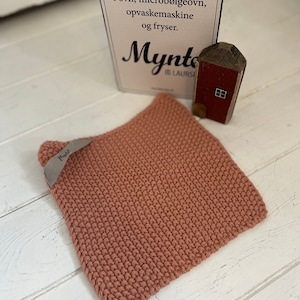 Peut inclure: Un dessous de plat en tricot rose avec le mot "Mynto" dessus. Le dessous de plat est posé sur une surface en bois blanc. Le texte sur la carte derrière le dessous de plat est le suivant : "Mynte kan benyttes i ovn, microbølgeovn, opvaskemaskine og fryser."