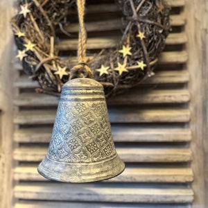 Könnte beinhalten: Ein rustikaler Zweigkranz mit goldenen Sternen hängt an einem Seil, das an einer silbernen Glocke mit dekorativem Muster befestigt ist. Die Glocke hängt an einem weißen Holzladen.