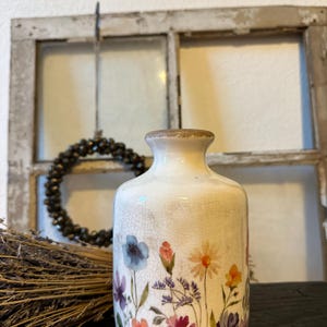 Könnte beinhalten: Eine weiße Keramikvase mit einem Blumendesign mit blauen, rosa und gelben Blumen. Die Vase hat ein leicht gealtertes, craquelé Aussehen und einen braunen Rand. Es ist ein dekorativer Einrichtungsgegenstand.