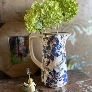 Puede incluir: Una jarra de cerámica con diseño floral en azul y rosa, con hortensias verdes. Una pequeña figura de un niño con una flor azul está frente a la jarra. La jarra tiene un asa y borde color crema.