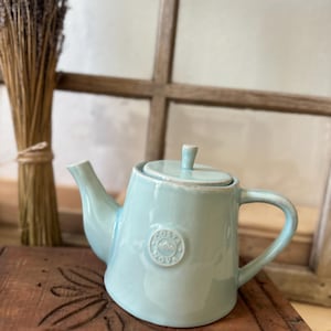 Può includere: Una teiera in ceramica blu chiaro con un coperchio e un manico. La teiera ha un logo rotondo sul lato che dice "Cotta Dove".