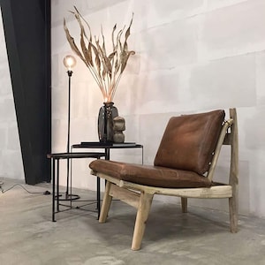 May include: A brown leather chair with a wooden frame and a curved back. The chair is sitting on a concrete floor in a room with white walls. There is a dried plant arrangement in a vase and a black floor lamp in the background.