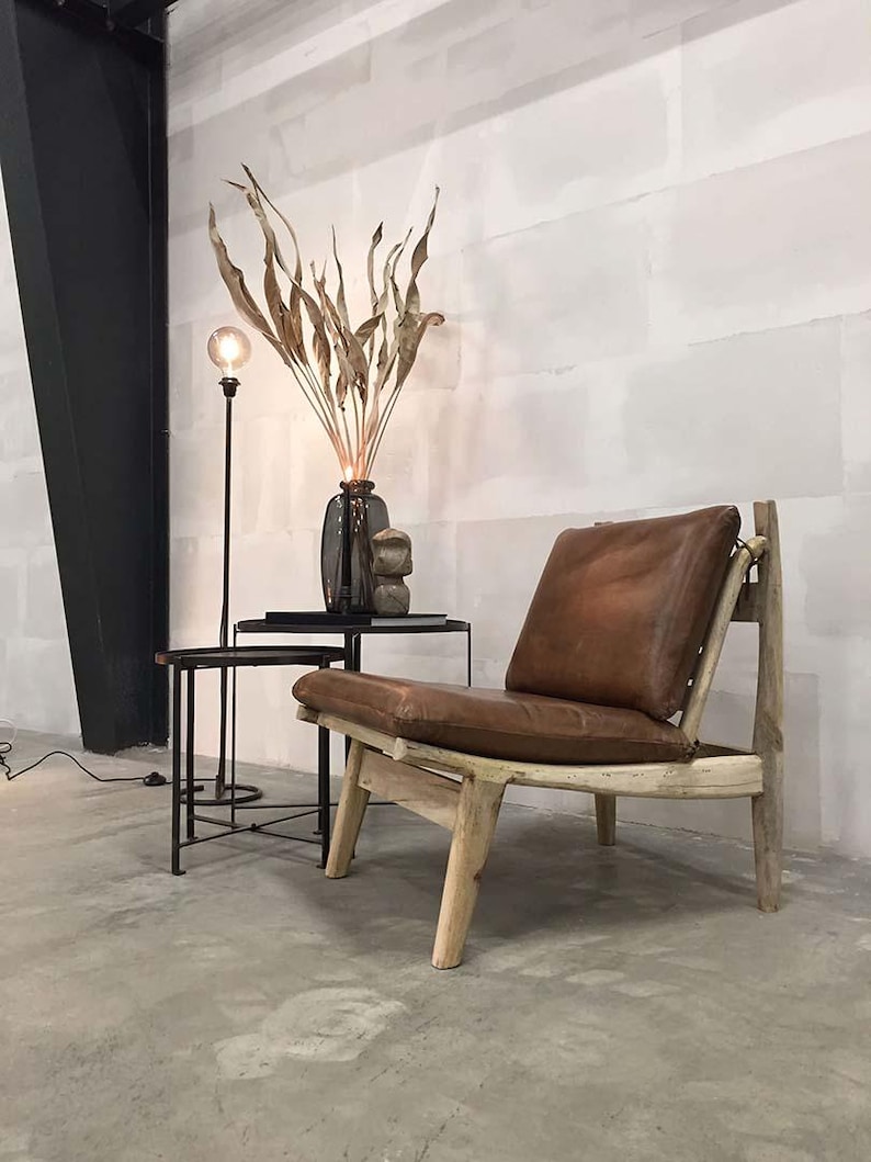 May include: A brown leather chair with a wooden frame and a curved back. The chair is sitting on a concrete floor in a room with white walls. There is a dried plant arrangement in a vase and a black floor lamp in the background.