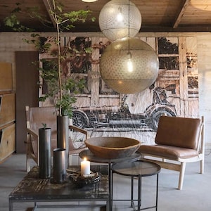 May include: A living room with a brown leather armchair, a wooden coffee table and two metal side tables. The coffee table has a wooden bowl on top. There are three hanging light fixtures with a geometric pattern. The walls are decorated with a large tapestry featuring a vintage car.