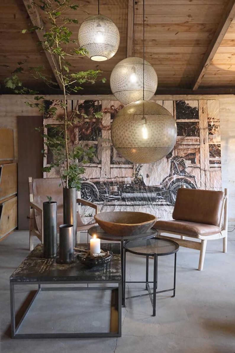May include: A living room with a brown leather armchair, a wooden coffee table and two metal side tables. The coffee table has a wooden bowl on top. There are three hanging light fixtures with a geometric pattern. The walls are decorated with a large tapestry featuring a vintage car.