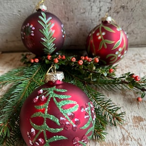 Op de afbeelding: Drie matte rode kerstballen met handgeschilderde groene botanische ontwerpen, waaronder dennennaalden en bessen. Elke bal heeft een gouden metalen dop en een gouden ophangkoord, geaccentueerd door natuurlijke groenblijvende takjes.