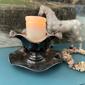 Puede incluir: Un candelero de plata con un diseño acanalado y una vela blanca en su interior. El candelero está sobre una superficie azul con una figurita de caballo blanco y una corona de flores secas en el fondo.