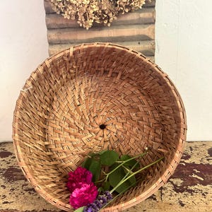 May include: A round, woven brown wicker basket with a spiral pattern and a small circular opening at the centre base. Inside the basket, there are two vibrant magenta flowers with green stems and a sprig of purple lavender with green leaves.