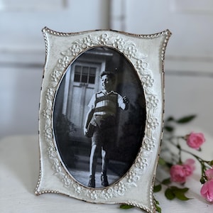 Könnte beinhalten: Ein cremefarbener, ovaler Bilderrahmen mit einem Schwarz-Weiß-Foto. Der Rahmen hat ein Blumendesign und einen Perlenrand. Er steht auf einer weißen Oberfläche, mit rosa Rosen im Hintergrund.