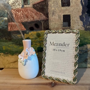 May include: A green and gold picture frame with a white mat and the text "Meander Elegant home decorations 10 x 15cm www.meander-bv.nl" on the front. The frame is sitting on a wooden surface next to a white ceramic vase with pink flowers.