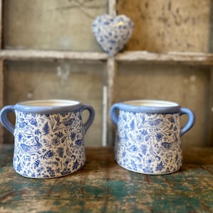 Könnte beinhalten: Zwei Keramikbecher mit blauen Griffen und Rändern. Die Becher haben einen weißen Hintergrund mit einem blauen Blumen- und Tiermuster. Eine blaue herzförmige Dekoration hängt im Hintergrund.