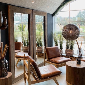 May include: Two brown leather chairs with wooden frames and a wooden coffee table in a room with large windows and a rustic wooden ceiling. The chairs are facing each other and the table is between them. There are plants in pots on the windowsill.