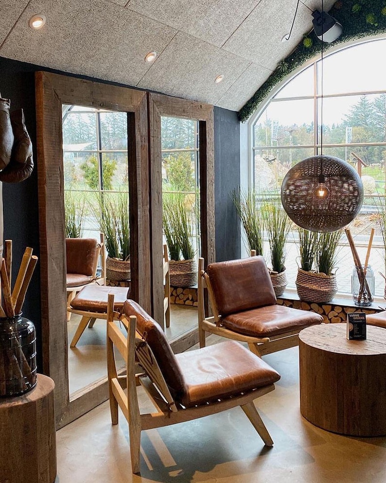 May include: Two brown leather chairs with wooden frames and a wooden coffee table in a room with large windows and a rustic wooden ceiling. The chairs are facing each other and the table is between them. There are plants in pots on the windowsill.