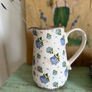 Peut inclure: Un pichet en céramique blanche avec un motif floral bleu. Le pichet a une anse et un bord légèrement évasé. Le pichet est posé sur une surface en bois vert.