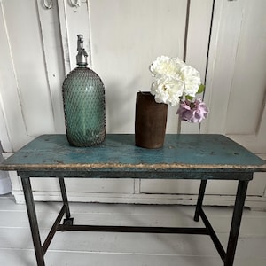 Könnte beinhalten: Ein rechteckiger Tisch mit einer abgenutzten, blau lackierten Oberfläche und dunklen Metallbeinen. Auf dem Tisch stehen ein grüner Glassodaspender mit Drahtgeflecht sowie eine dunkelbraune zylindrische Vase mit weißen und hellvioletten Blumen.